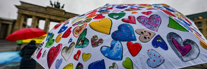 Das Brandenburger Tor steht im Hintergrund, davor halten Menschen einen bunten Regenschirm voller Herzen. Es regnet, aber die Farben des Schirms leuchten fröhlich im grauen Wetter.