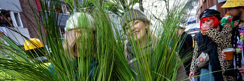 Zwei als Grasbüschel verkleidete Frauen stehen am Rand des Zugwegs des Rosenmontagszugs.