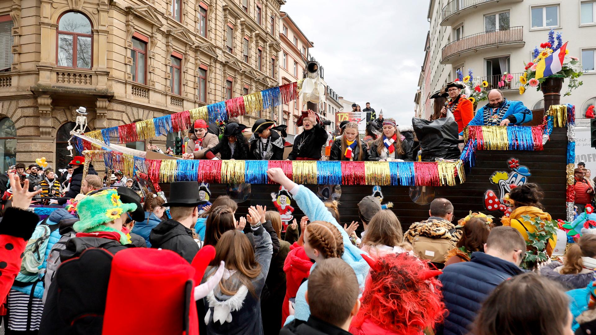 Karnevalswagen, der als Piratenschiff geschmückt ist. Darauf sind Menschen zu sehen, die als Piraten verkleidet sind. Vor dem Wagen stehen verkleidete Menschen.