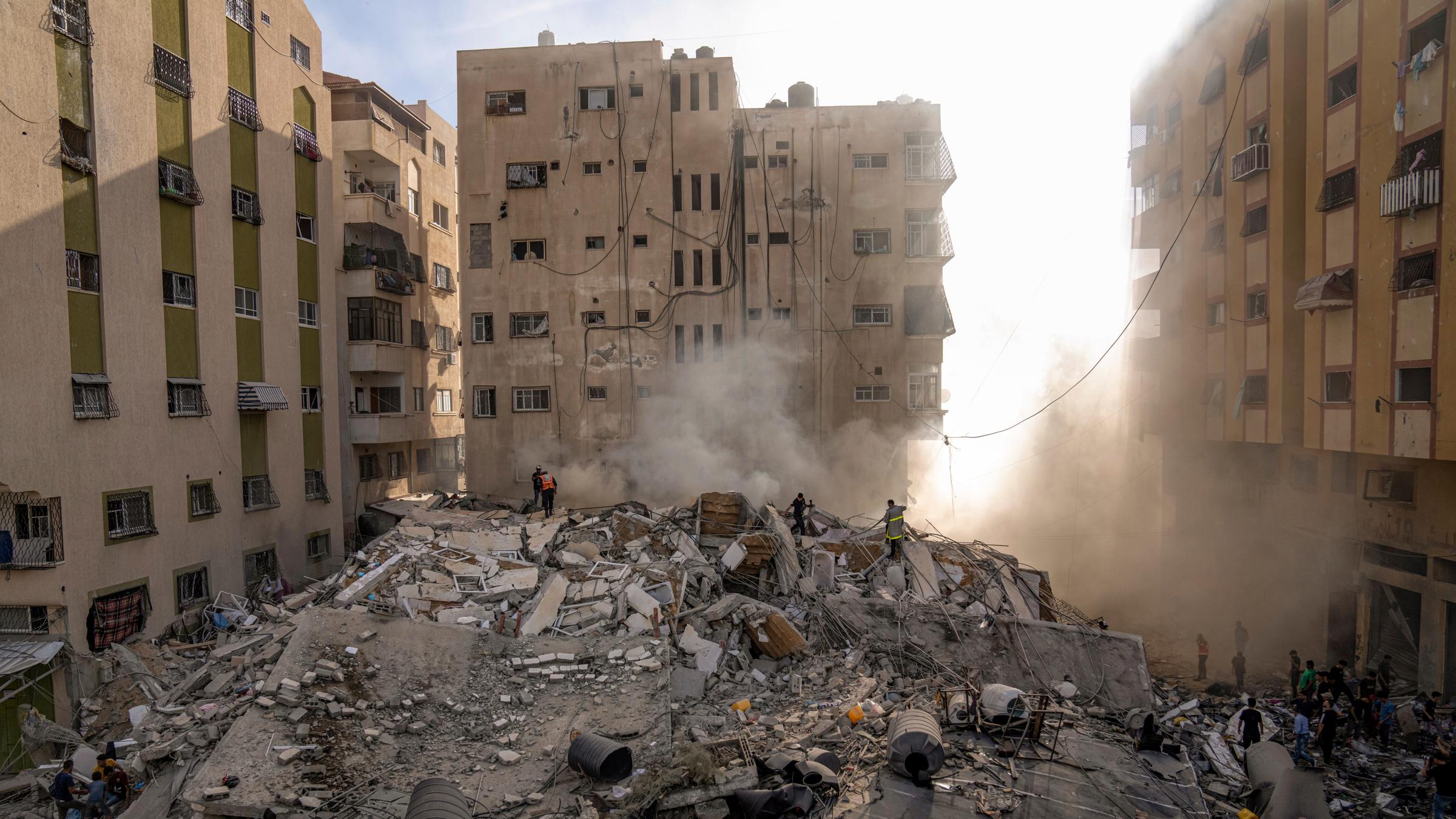Drei große, halb zerstörte Häuser rahmen einen Platz ein, auf dem Trümmer und Geröll liegen, im Hintergrund staubt es.