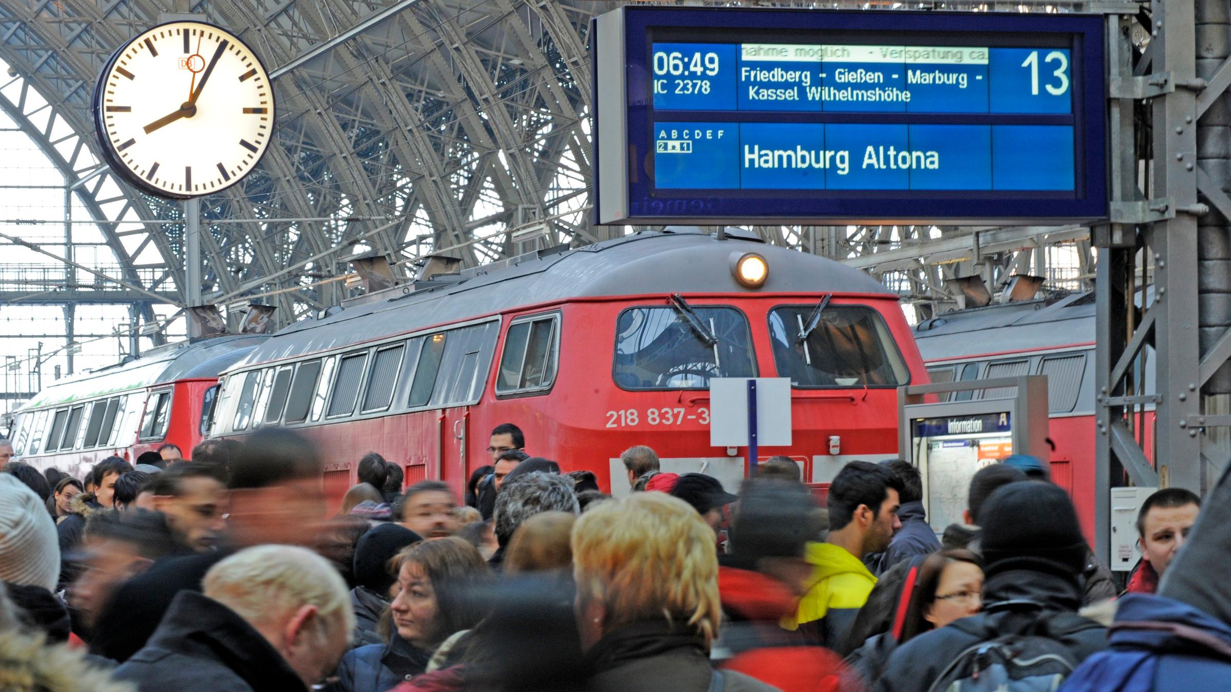 Viele Menschen stehen auf einem Bahnsteig, auf dem Gleis ein roter Zug. Oben eine Anzeigetafel und eine Bahnhofsuhr.