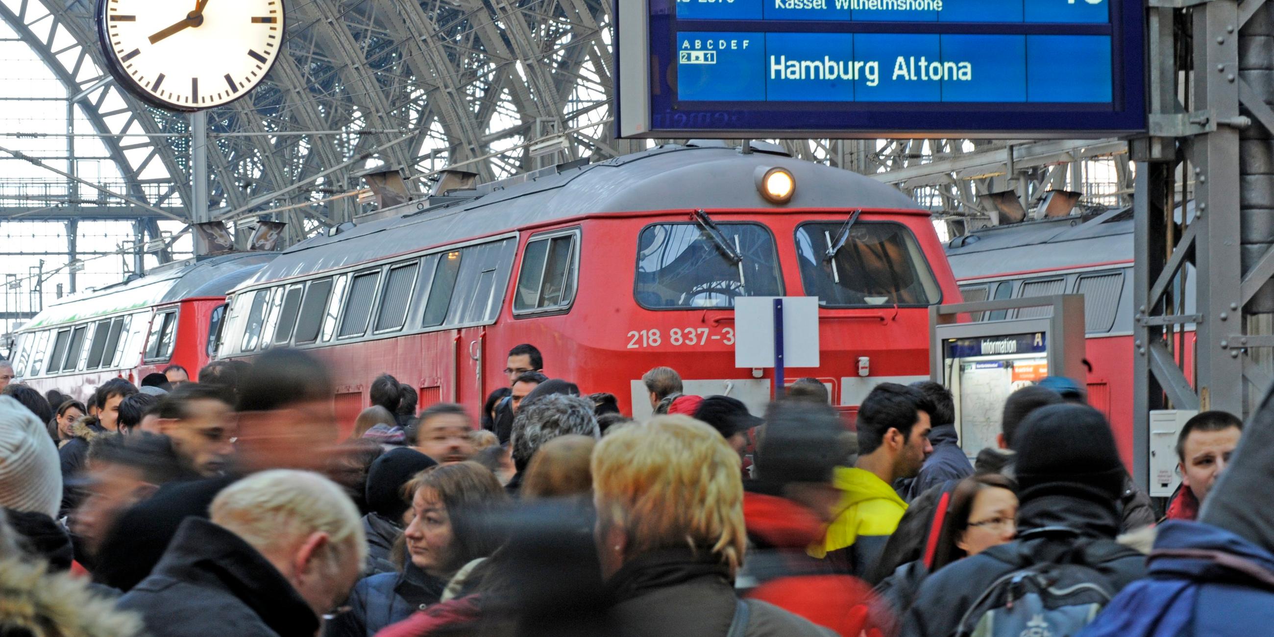 Viele Menschen stehen auf einem Bahnsteig, auf dem Gleis ein roter Zug. Oben eine Anzeigetafel und eine Bahnhofsuhr.