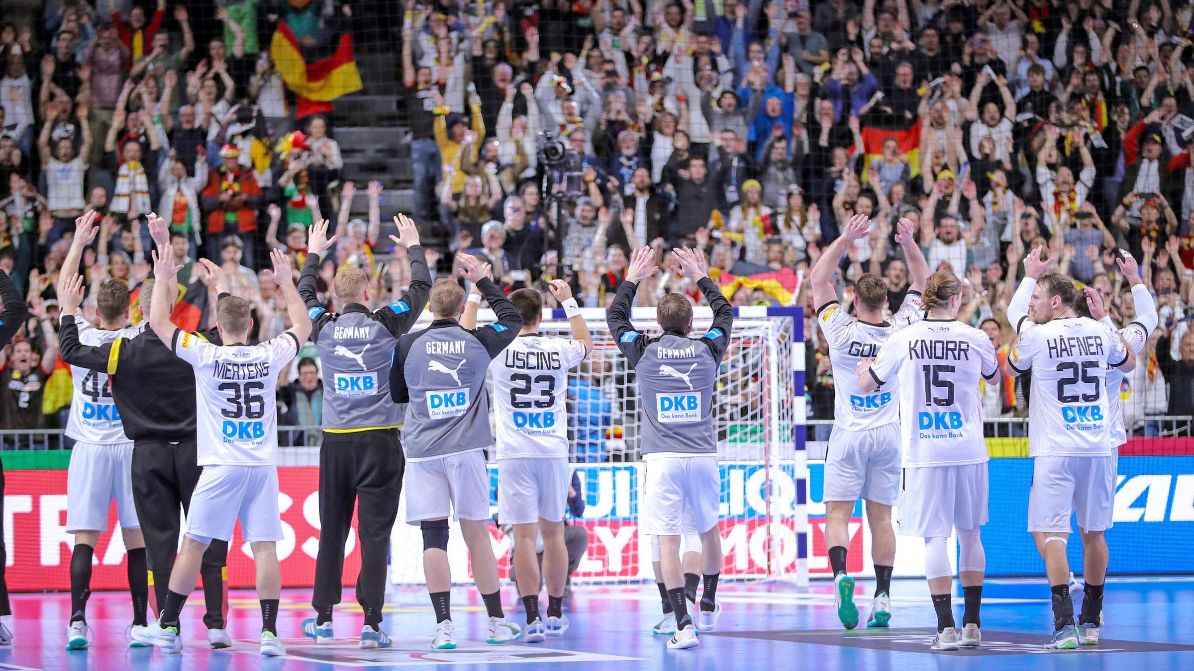 Handballer stehen auf dem Spielfeld mit dem Rücken zur Kamrea und jubeln ihren Fans zu. Im Hintergund voll Tribünen und das Tor.