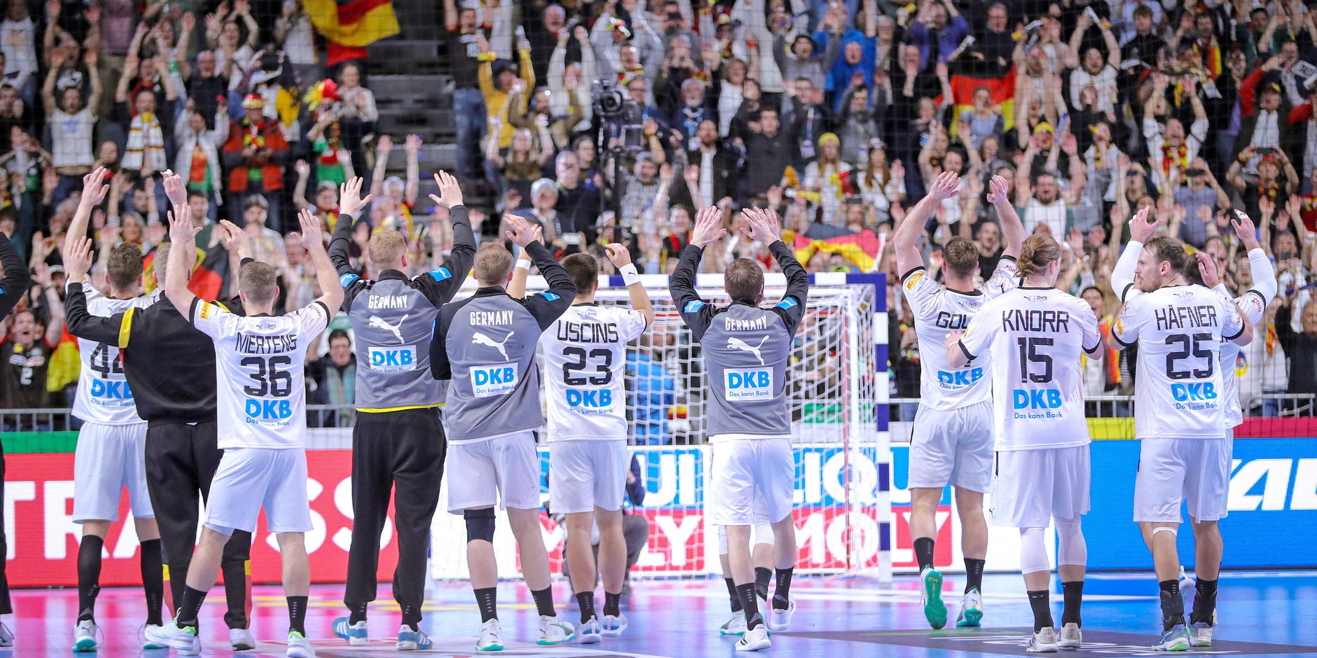 Handballer stehen auf dem Spielfeld mit dem Rücken zur Kamrea und jubeln ihren Fans zu. Im Hintergund voll Tribünen und das Tor.