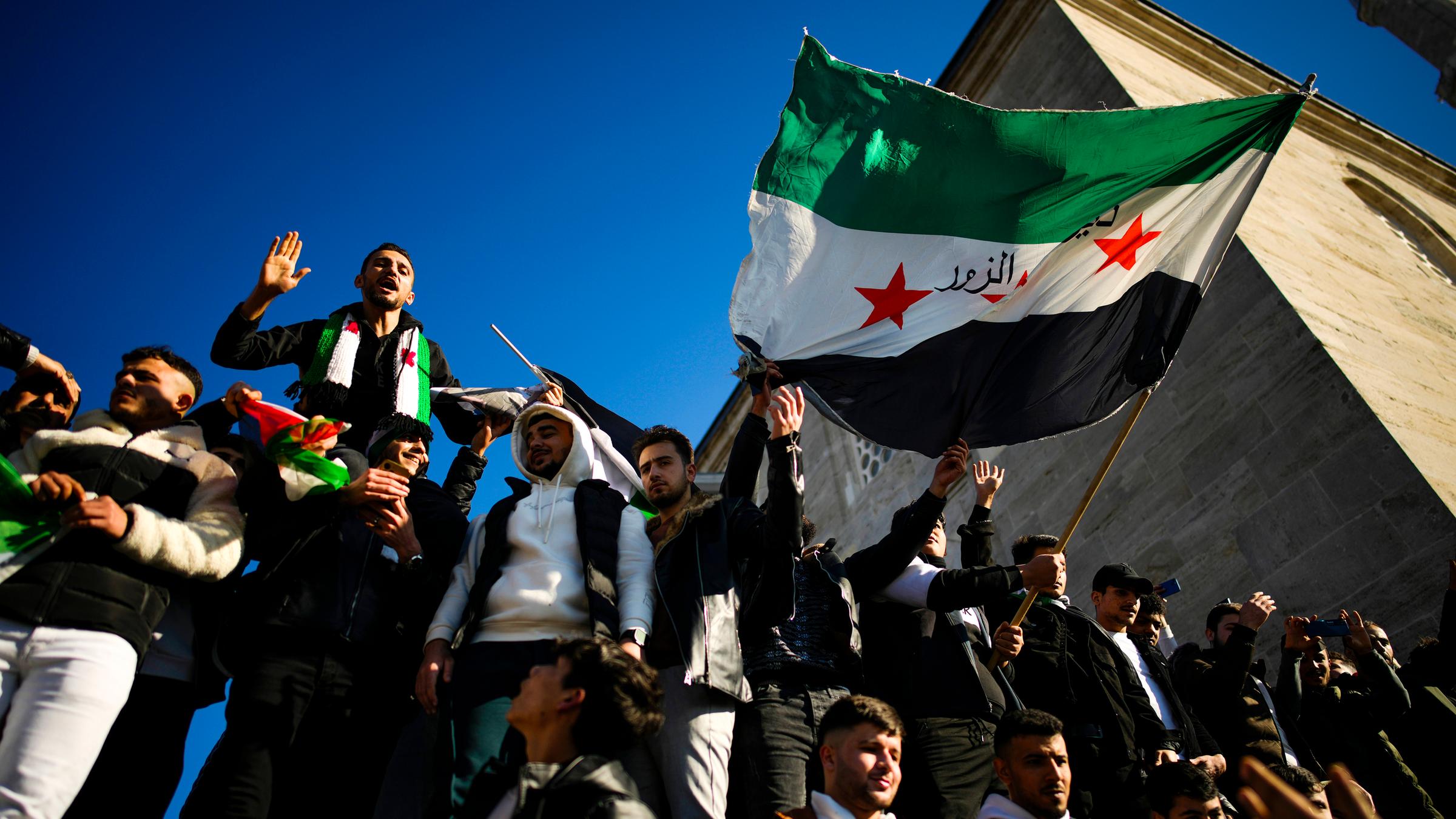 Syrische Menschen feiern, halten die syrische Flagge in die Luft. Im Hintergund blauer Himmel und der Turm einer Moschee.