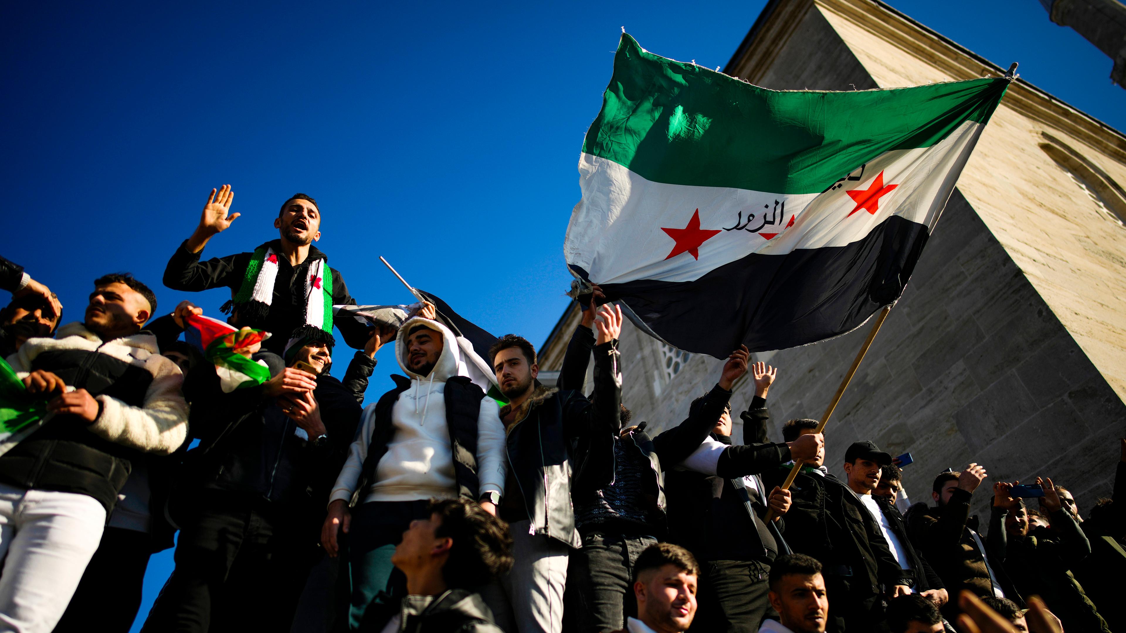 Syrische Menschen feiern, halten die syrische Flagge in die Luft. Im Hintergund blauer Himmel und der Turm einer Moschee.