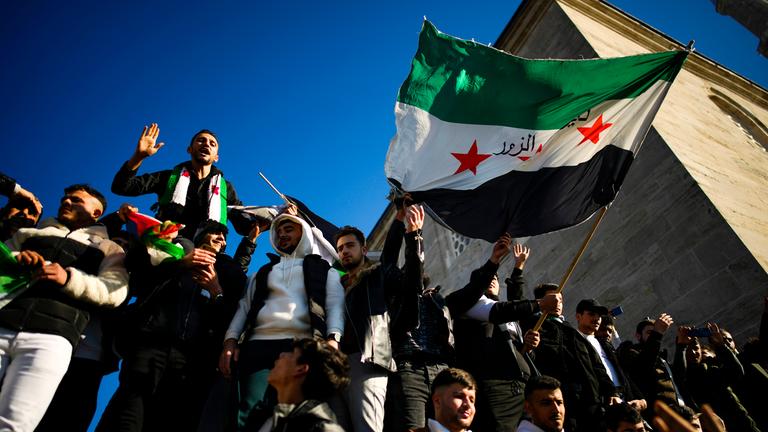 Syrische Menschen feiern, halten die syrische Flagge in die Luft. Im Hintergund blauer Himmel und der Turm einer Moschee.