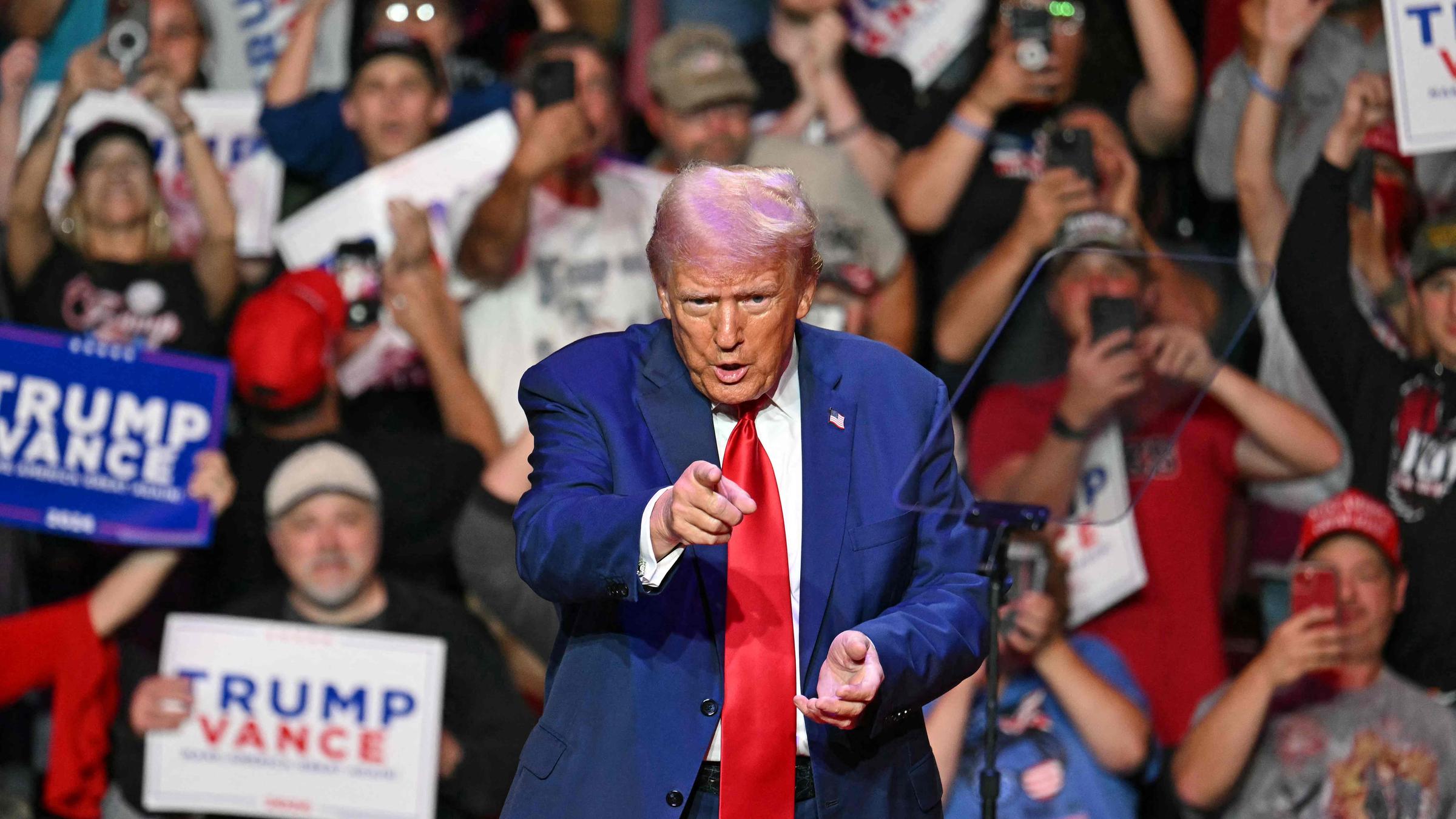 Donald Trump zeigt mit dem Zeigenfinger geradeaus. Im Hintergrund viele Menschen, die filmen, jubeln und Transparente halten.