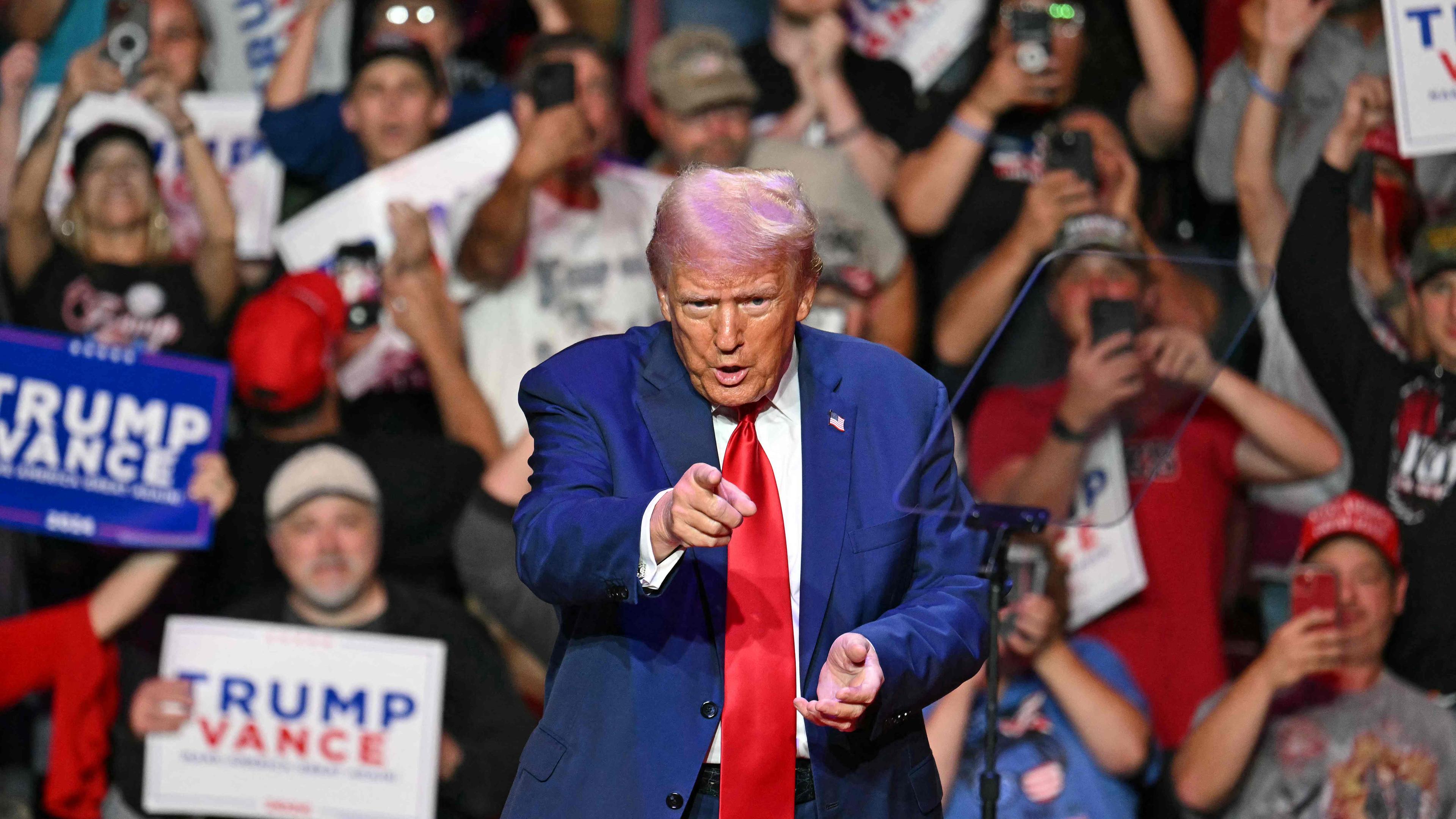 Donald Trump zeigt mit dem Zeigenfinger geradeaus. Im Hintergrund viele Menschen, die filmen, jubeln und Transparente halten.