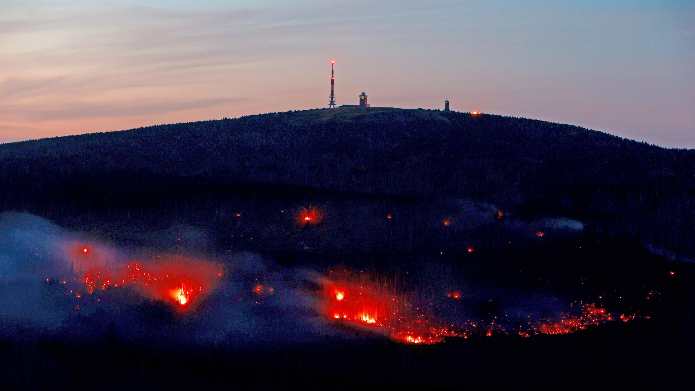 Ein dunkler Berg mit hell leuchtenden Glutnestern. Au dem Berggipferl stehen Masten. Im Hintergrund orangener Himmel durch die untergehende Sonne.
