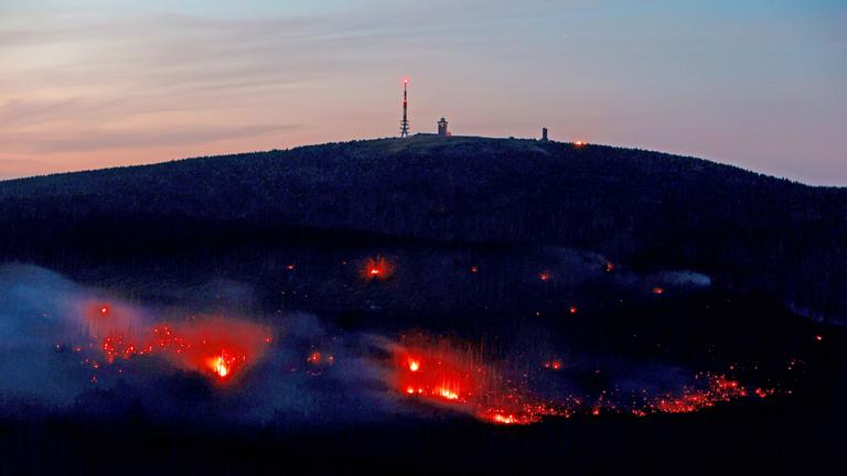 Ein dunkler Berg mit hell leuchtenden Glutnestern. Au dem Berggipferl stehen Masten. Im Hintergrund orangener Himmel durch die untergehende Sonne.