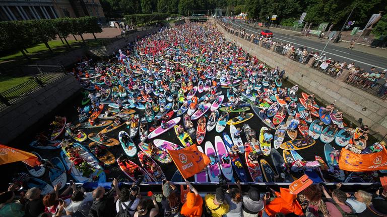 Bei einem Festival in St. Petersburg, Russland, paddeln viele Menschen auf ihren Stand-up-Paddle-Boards über einen Fluss. Und schaut mal genauer hin: Viele haben sich dafür richtig kreativ verkleidet! 😄