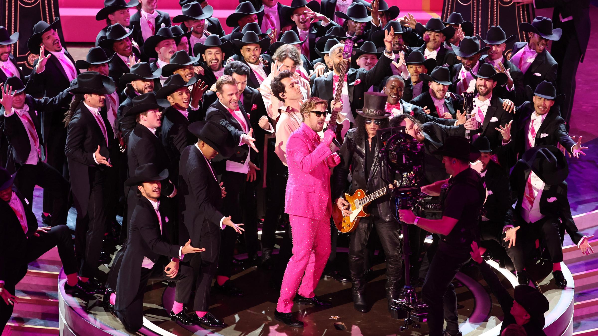 Ryan Gosling sing im pinken Glitzeranzug. Um ihn herum stehen 56 in schwarz gekleidete Tänzer.