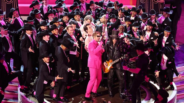 Ryan Gosling sing im pinken Glitzeranzug. Um ihn herum stehen 56 in schwarz gekleidete Tänzer.