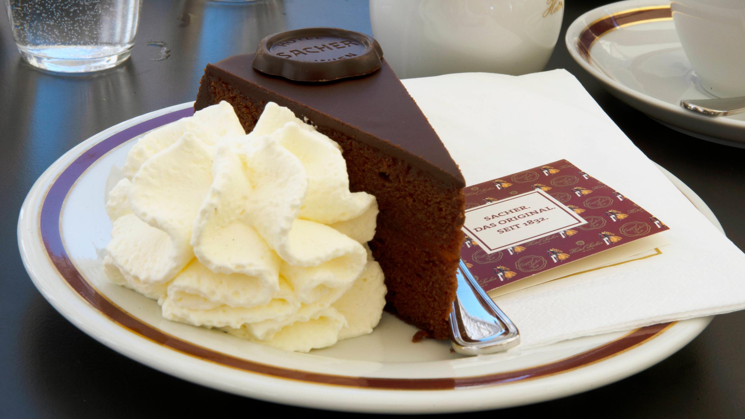 Auf einem Kuchenteller steht ein Stück Sachertorte mit Schokolade und Sahne. 