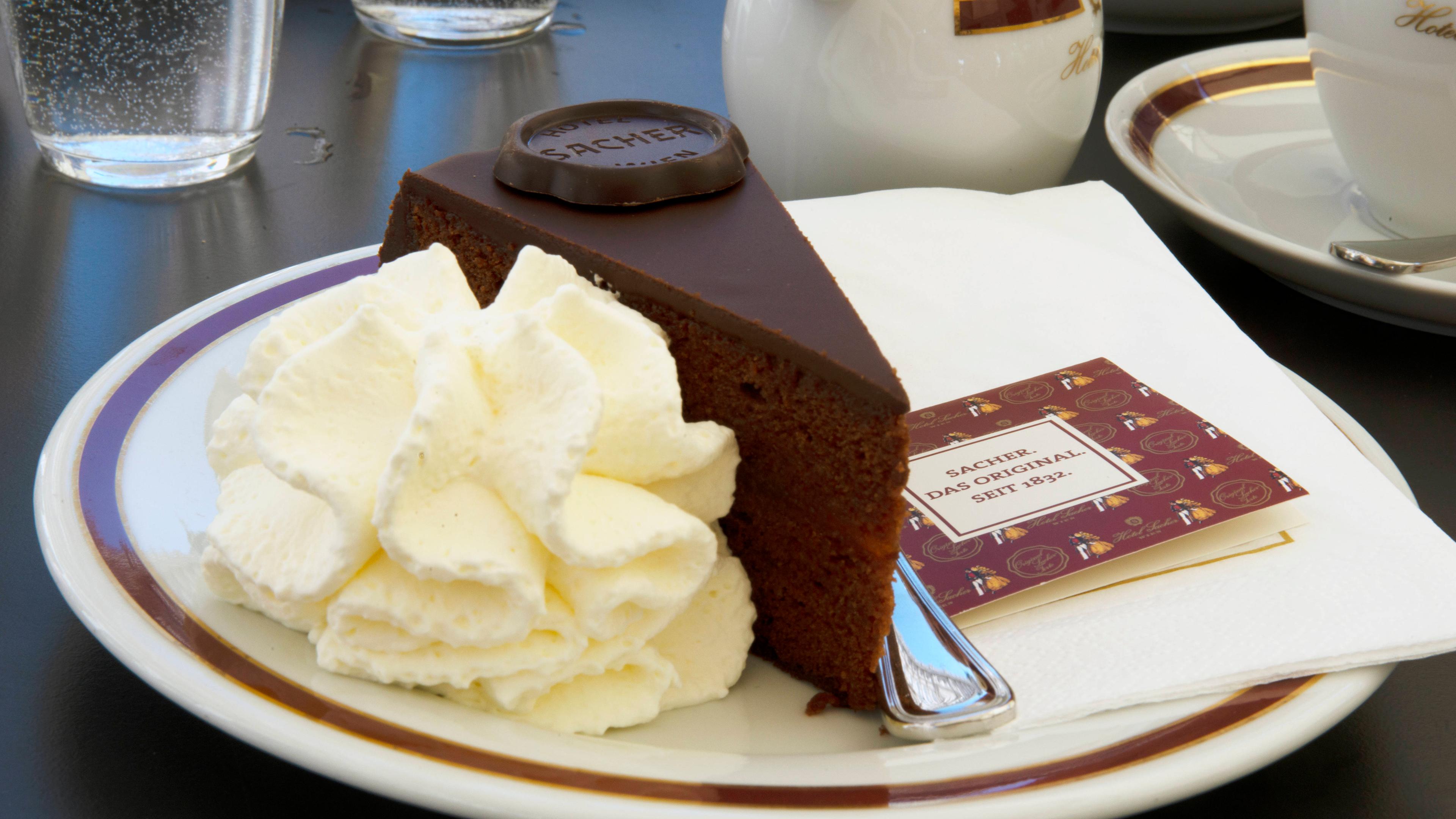 Auf einem Kuchenteller steht ein Stück Sachertorte mit Schokolade und Sahne. 