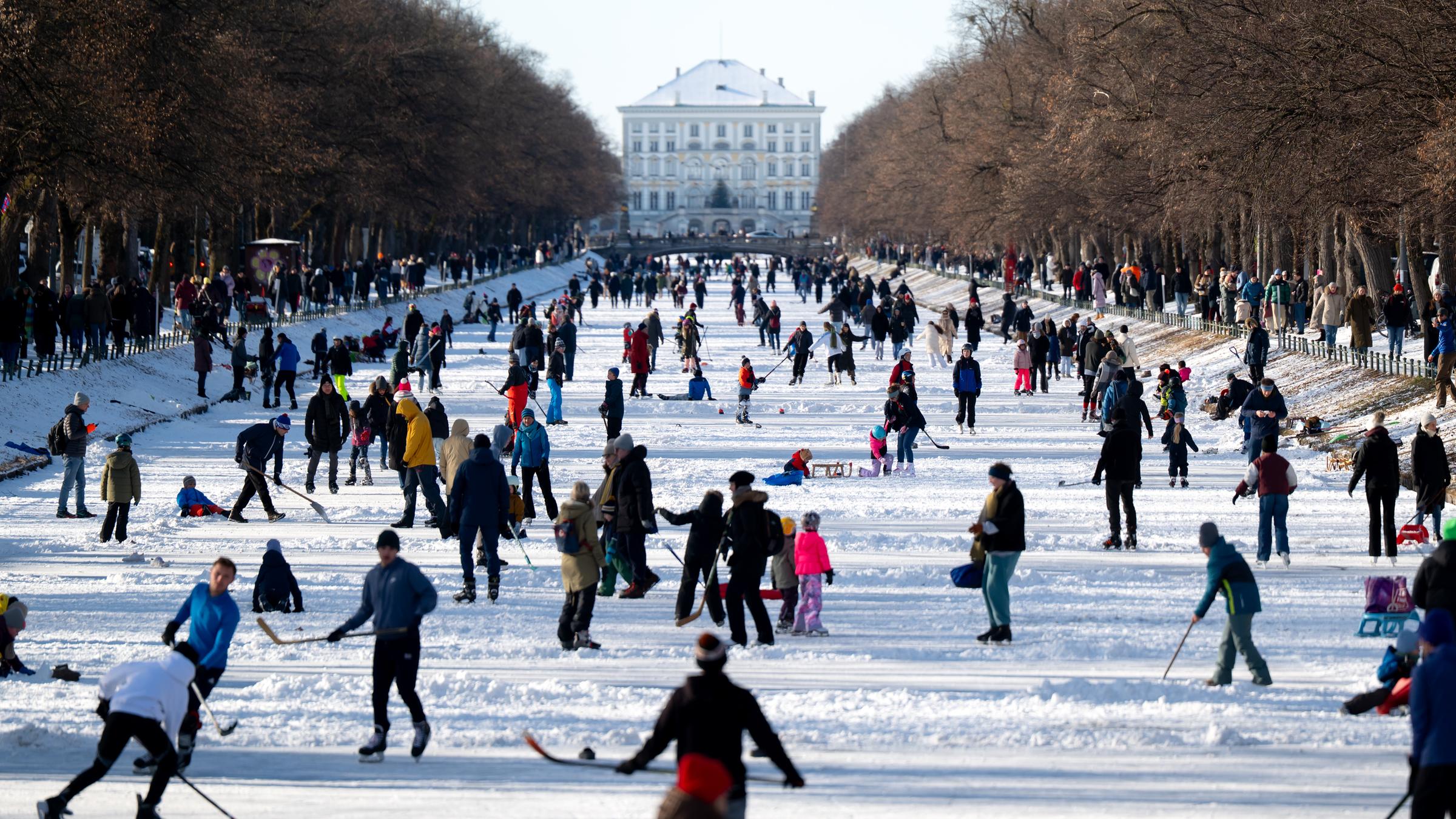Viele Menschen im Schnee vor dem Nymphenburger Schloss. Sie sind dick angezogen. Manche spazieren, andere spielen Eishockey auf dem zugefrorenen Nymphenburger Kanal in München.