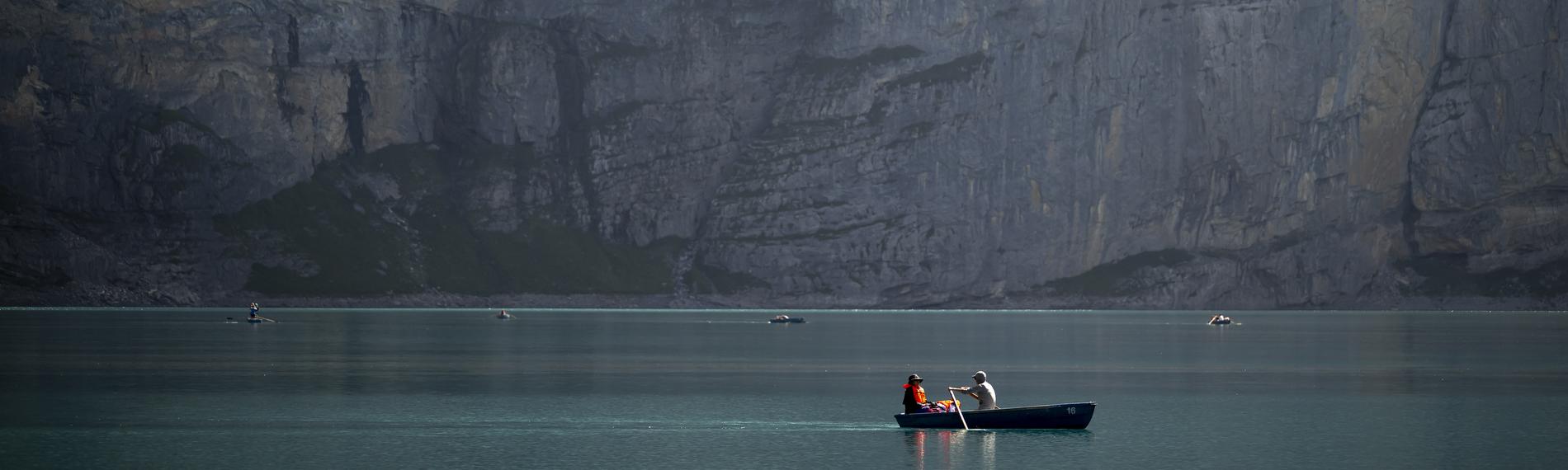 Ein kleines Ruderboot mit zwei Menschen auf einem See vor einer riesigen Felswand eines Bergs.