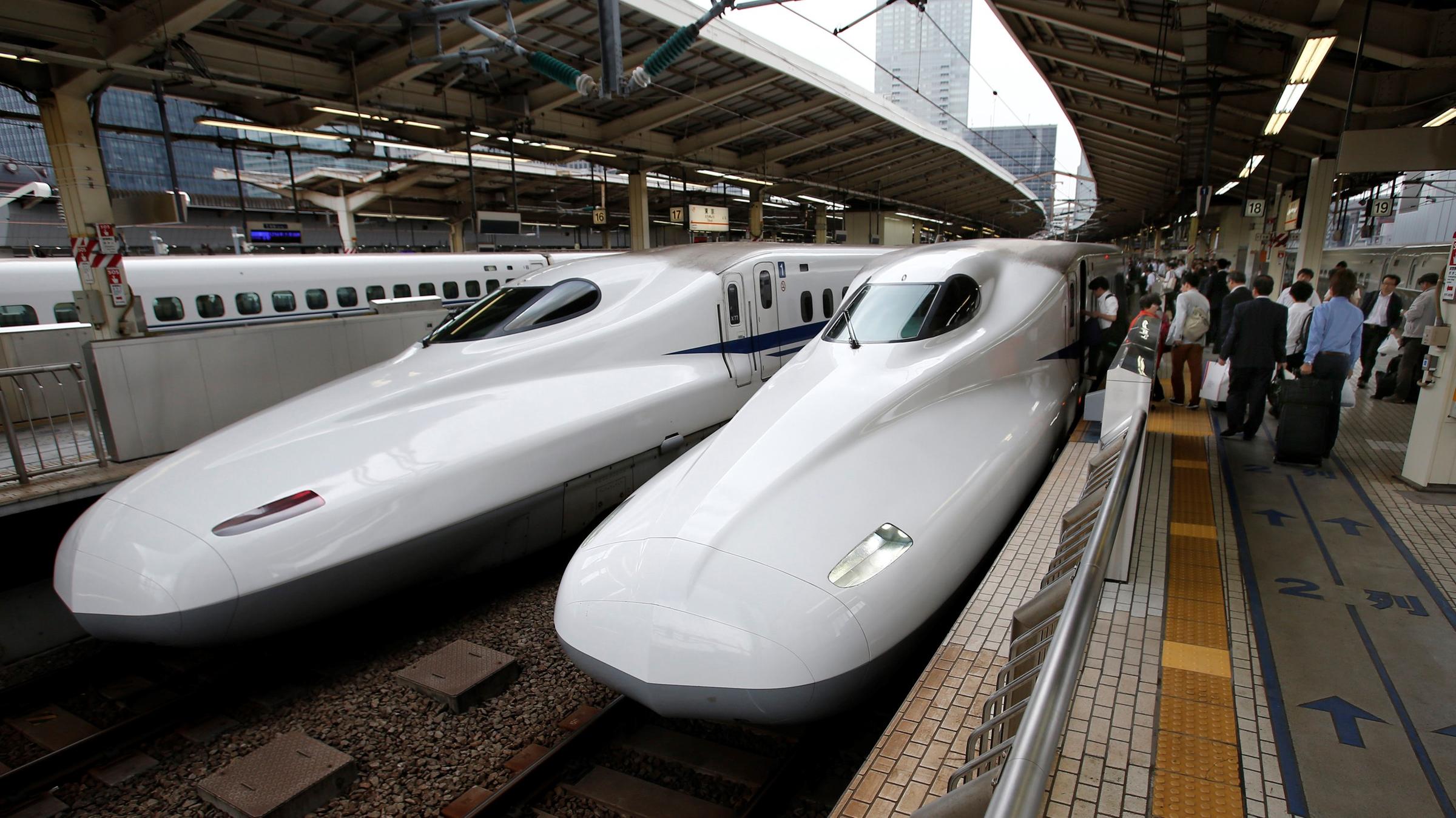 Zwei Shinkansen-Züge stehen in einem Bahnhof nebeneinander. Auf der rechten Seite steigen Menschen ein. Beide Züge sind weiß und haben eine lange Schnauze.