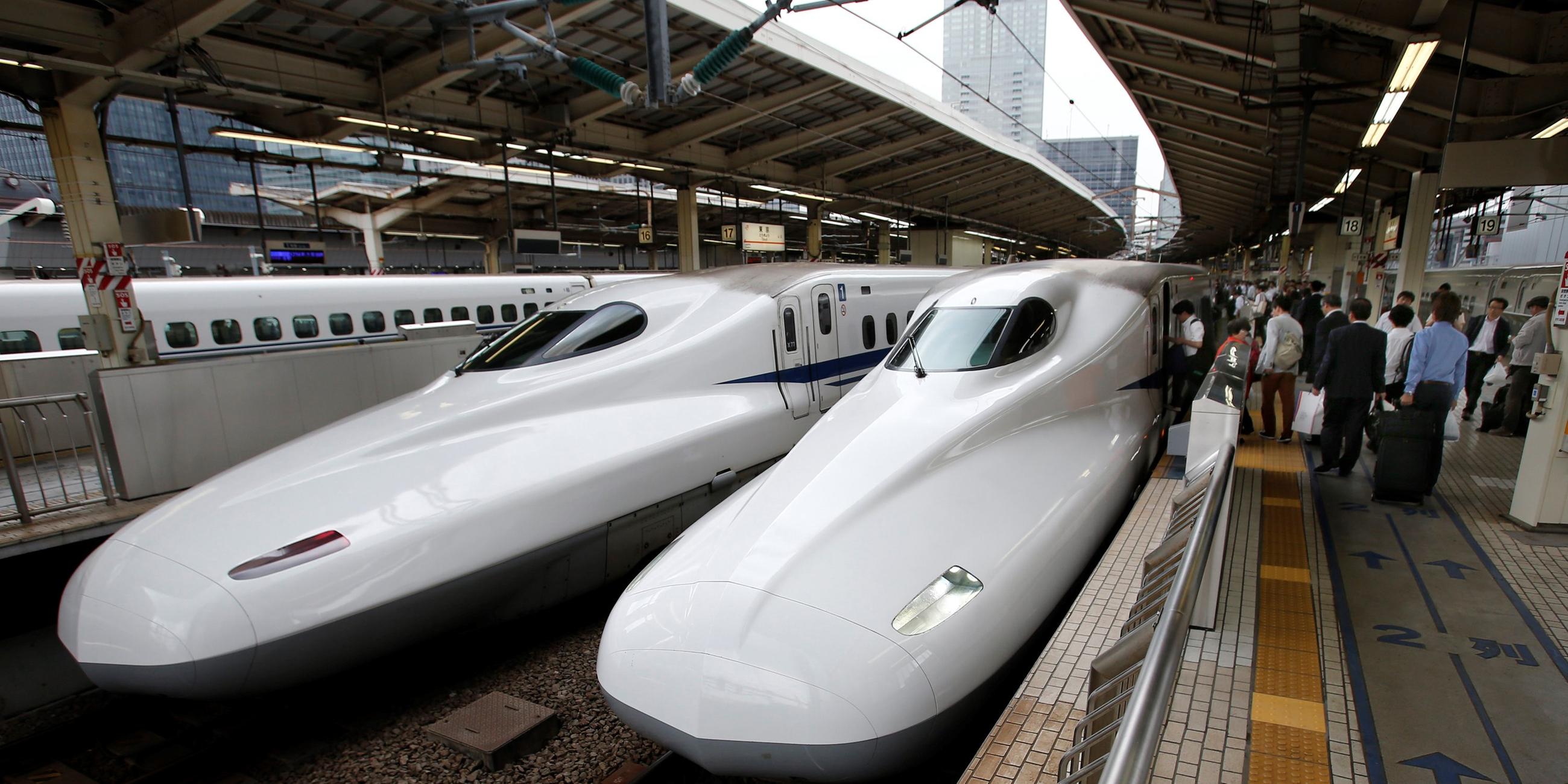 Zwei Shinkansen-Züge stehen in einem Bahnhof nebeneinander. Auf der rechten Seite steigen Menschen ein. Beide Züge sind weiß und haben eine lange Schnauze.