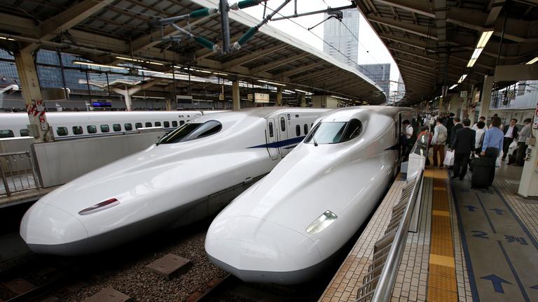 Zwei Shinkansen-Züge stehen in einem Bahnhof nebeneinander. Auf der rechten Seite steigen Menschen ein. Beide Züge sind weiß und haben eine lange Schnauze.