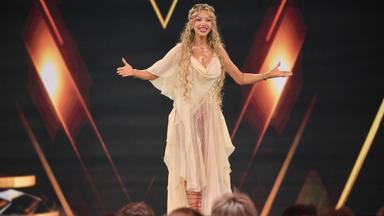 Grafik: Shirin David steht mit gehobenen Armen vor einem Publikum auf einer Bühne.