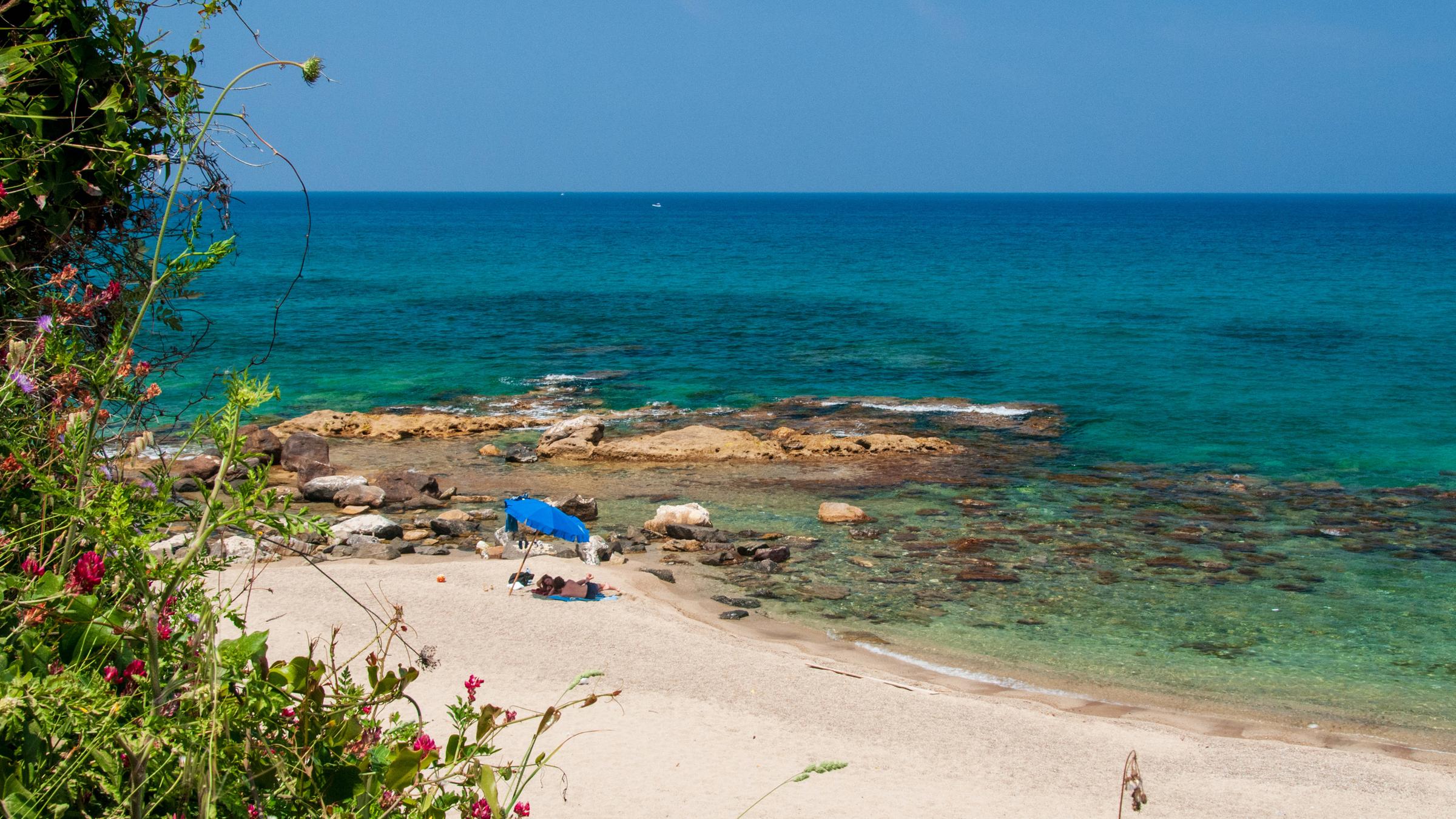 Man sieht einen Strand, an dem zwei Menschen unter einem blauen Sonnenschirm liegen. Das Wasser ist türkis und der Himmel blau.