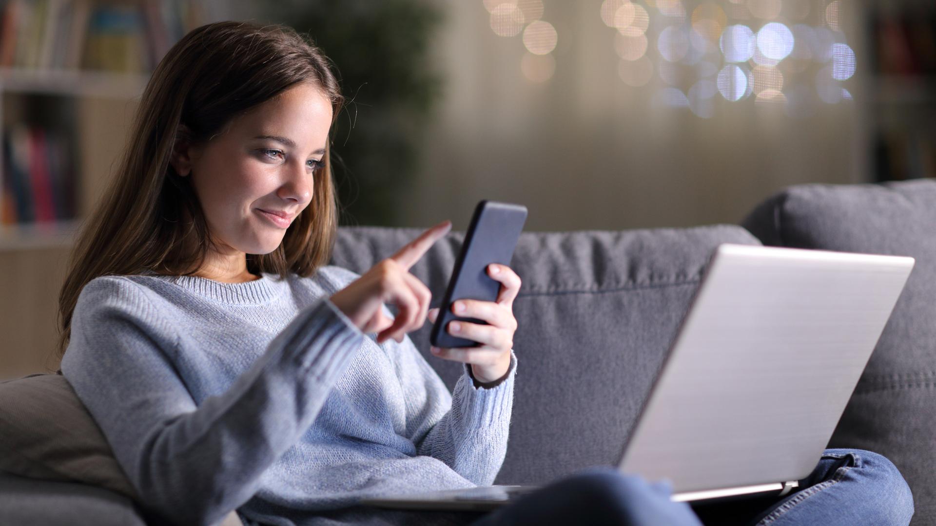 Auf dem Bild sieht man eine junge Frau im Schneidersitz auf einer Couch im Wohnzimmer sitzen. Auf ihrem Schoß hat sie einen Laptop und in der Hand einen Hand hält sie ein Smartphone. Ihr lächelnder Blick ist auf das Smartphone gerichtet - mit der anderen Hand wischt sie mit den Fingern über das Handydisplay.