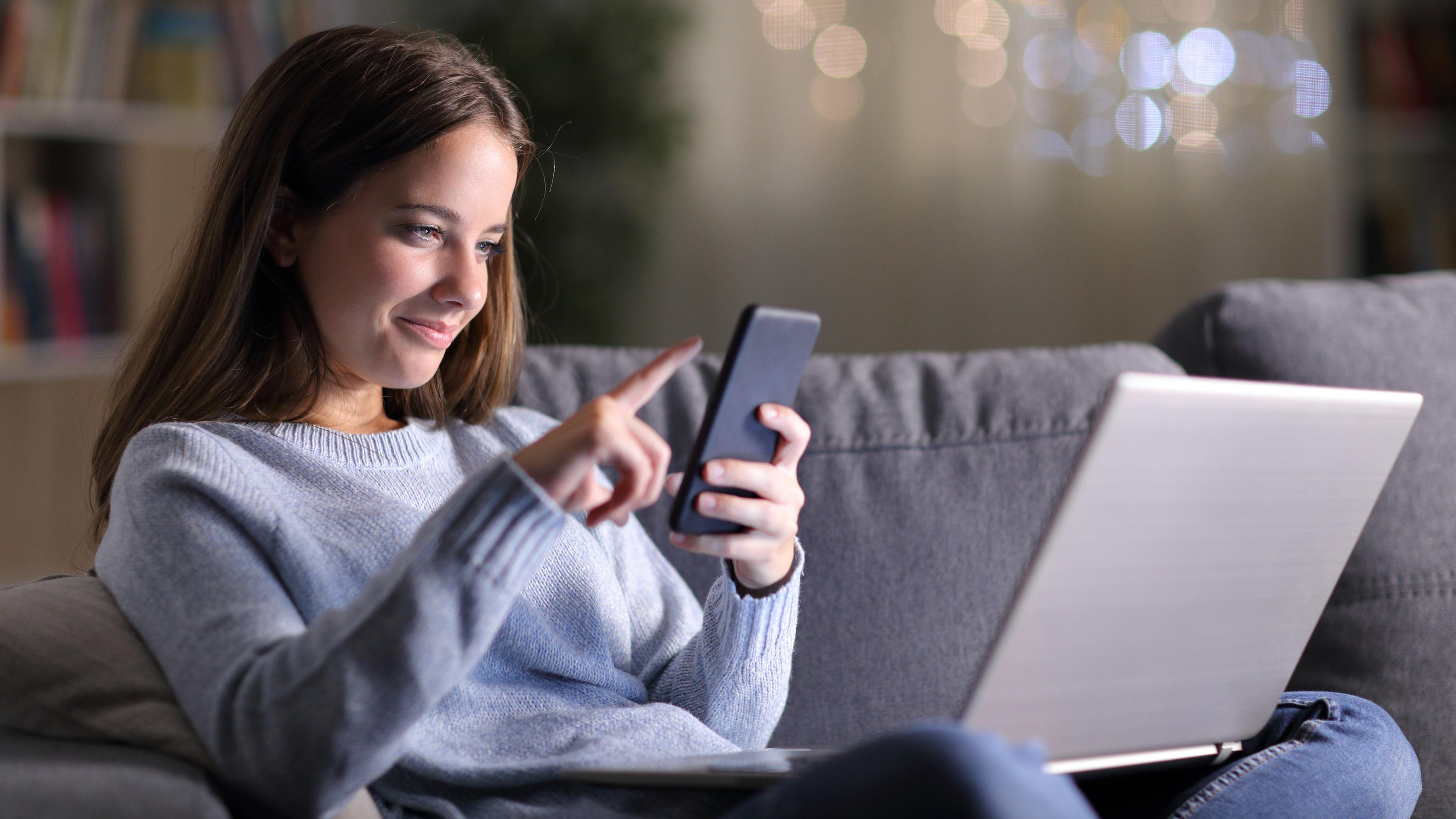 Auf dem Bild sieht man eine junge Frau im Schneidersitz auf einer Couch im Wohnzimmer sitzen. Auf ihrem Schoß hat sie einen Laptop und in der Hand einen Hand hält sie ein Smartphone. Ihr lächelnder Blick ist auf das Smartphone gerichtet - mit der anderen Hand wischt sie mit den Fingern über das Handydisplay.