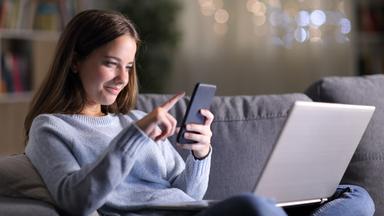 Auf dem Bild sieht man eine junge Frau im Schneidersitz auf einer Couch im Wohnzimmer sitzen. Auf ihrem Schoß hat sie einen Laptop und in der Hand einen Hand hält sie ein Smartphone. Ihr lächelnder Blick ist auf das Smartphone gerichtet - mit der anderen Hand wischt sie mit den Fingern über das Handydisplay.