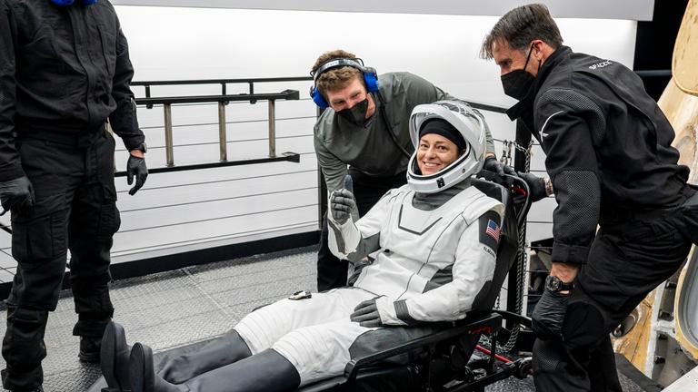 NASA-Astronautin Nicole Mann streckt den Daumen nach oben. Sie wird von mehreren SpaceX-Mitarbeitern auf einer Trage sitzend aus einer Raumkapsel geholt.