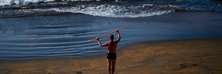 Ein Mann steht am Strand und macht Sportübungen mit einem Stock. 