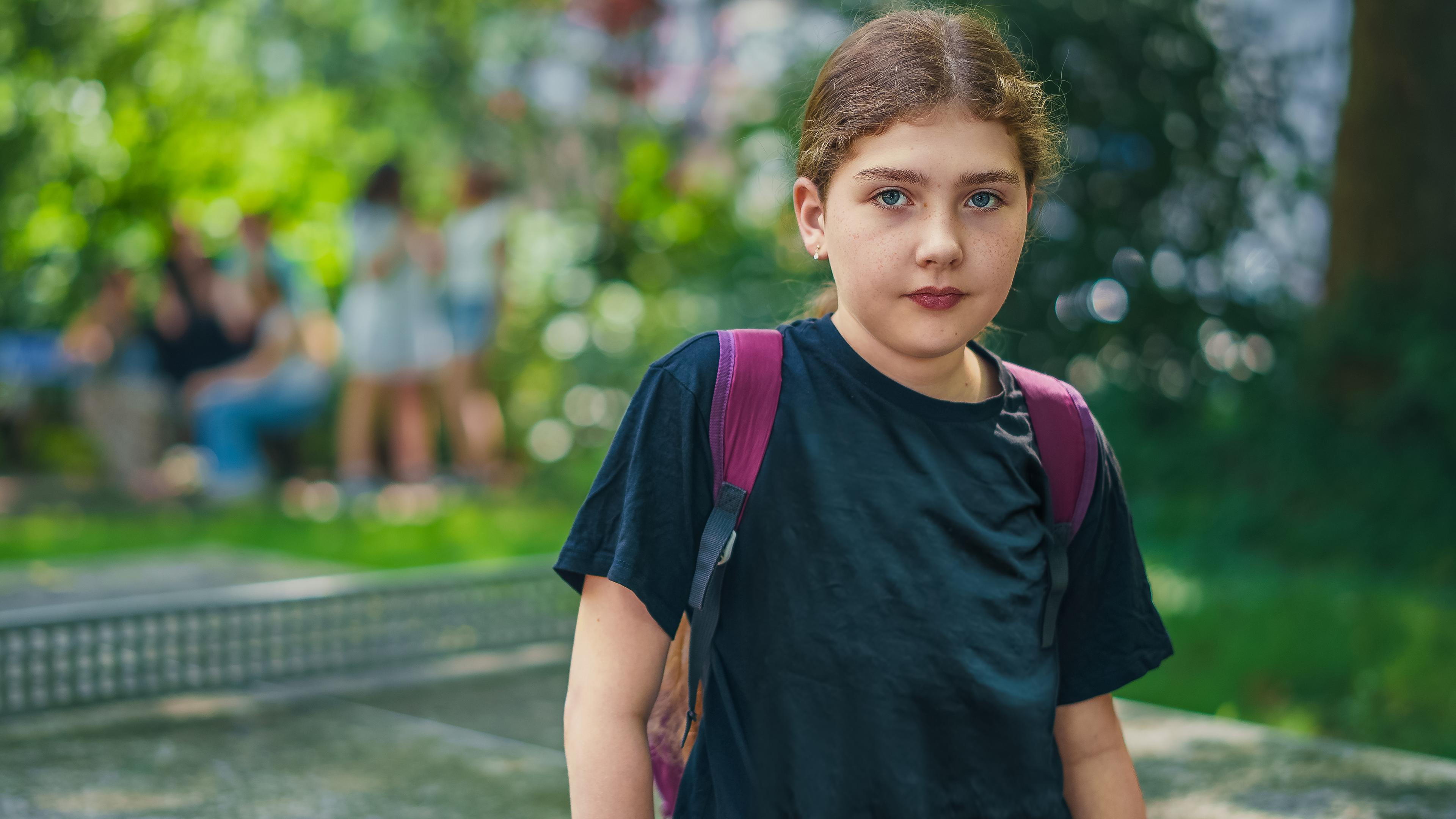 Lya (11) trägt ein schwarzes T-Shirt und einen lila Rucksack. Sie schaut ernst in die Kamera.