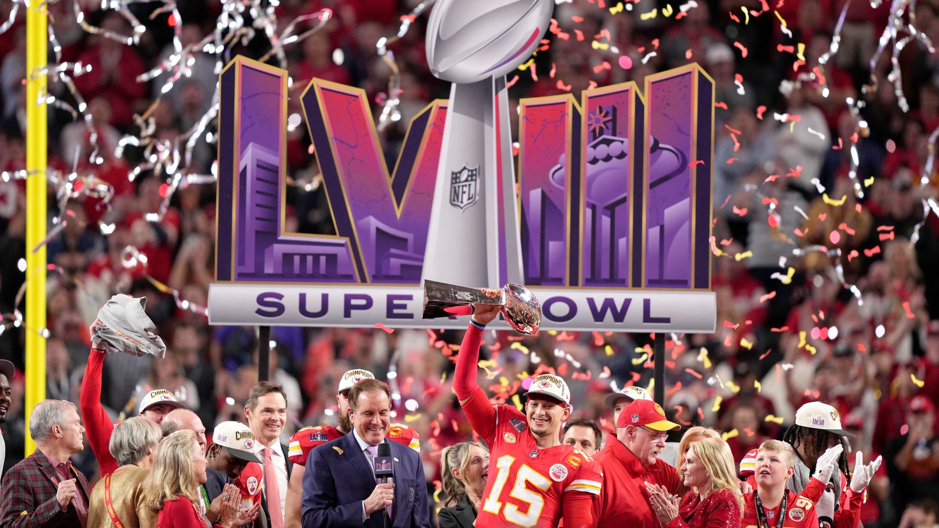 Patrick Mahomes hält die Super Bowl Trophäe in die Höhe, dahinter Konfetti, andere Menschen und das Logo des Super Bowls.