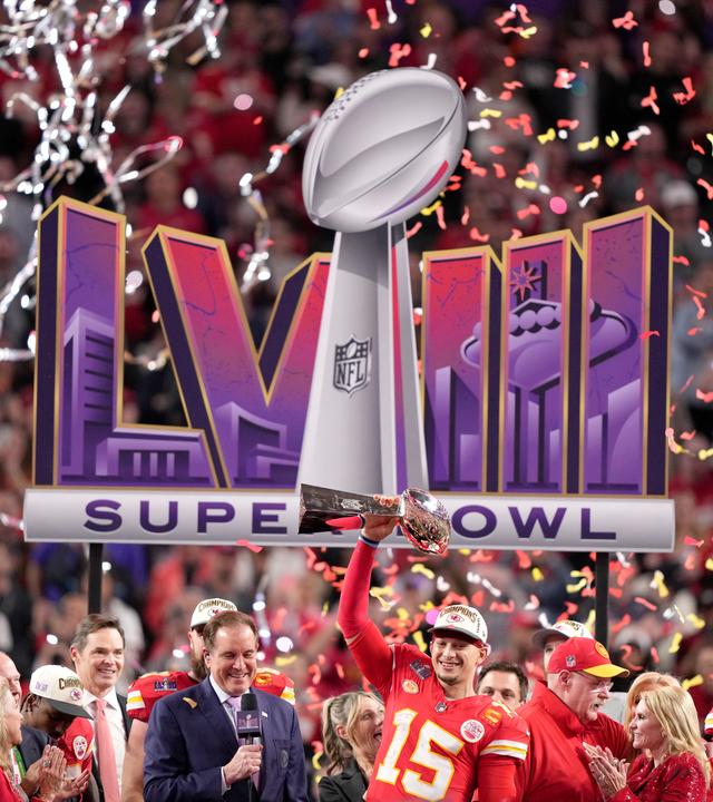 Patrick Mahomes hält die Super Bowl Trophäe in die Höhe, dahinter Konfetti, andere Menschen und das Logo des Super Bowls.