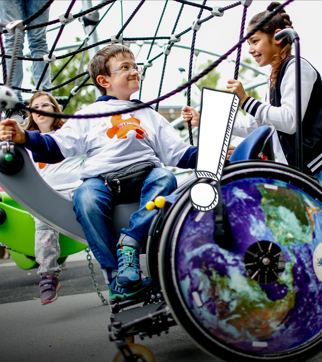 Ein Junge und ein Mädchen auf einem Spielplatz, davor steht ein Rollstuhl.