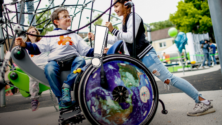 Ein Junge und ein Mädchen auf einem Spielplatz, davor steht ein Rollstuhl.