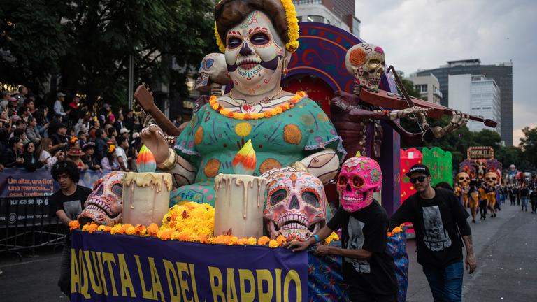 Ein großer Festwagen mit einer meterhohen weiblichen Totenkopf-Puppe und vielen weiteren bunten Totenköpfen als Dekoration zieht eine Parade entlang.