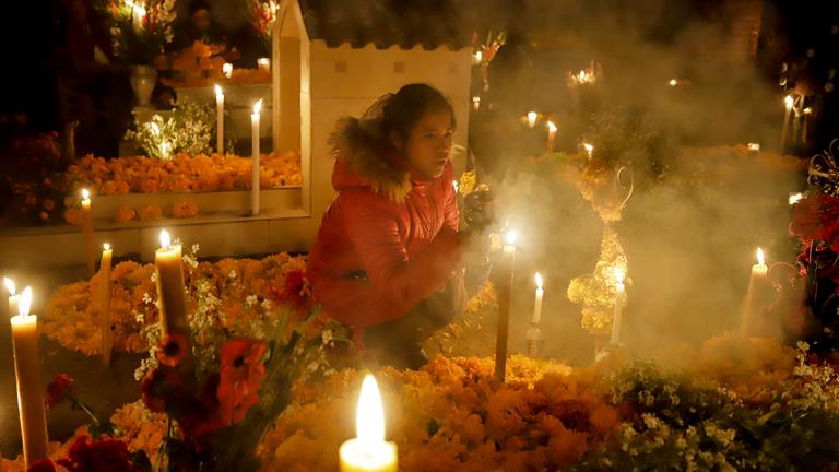Eine junge Frau sitzt in der Hocke zwischen blumenüberhäuften Gräbern. Es ist Nacht und die Kerzen, die auf den Gräbern stehen, leuchten hell und sondern hellen Rauch ab.