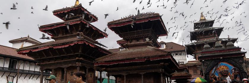 Menschen spazieren über den historischen Durbar Square. In der Luft fliegen Tauben. 