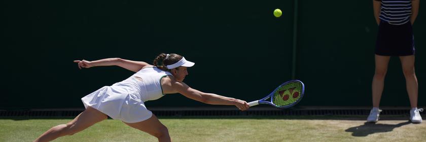 Elise Mertens trägt weiße Tenniskleidung und versucht, den Ball mit ausgestrecktem Arm zu erreichen. Gespielt wird auf einem grünen Tennisrasen.