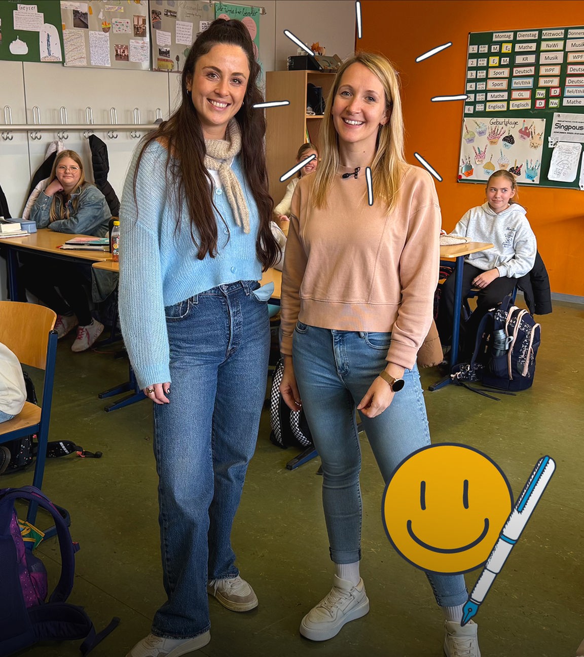 Reporterin Teresa steht neben Frau Jücker, eine Lehrerin an der IGS in Nieder-Olm. Sie stehen in einer Klasse, in der Kinder an Tischen sitzen und in die Kamera lächeln.