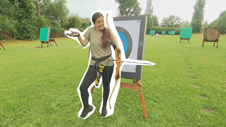 Teresa mit Pfeil und Bogen vor einer Zielscheibe - sie hebt unsicher die Arme. 