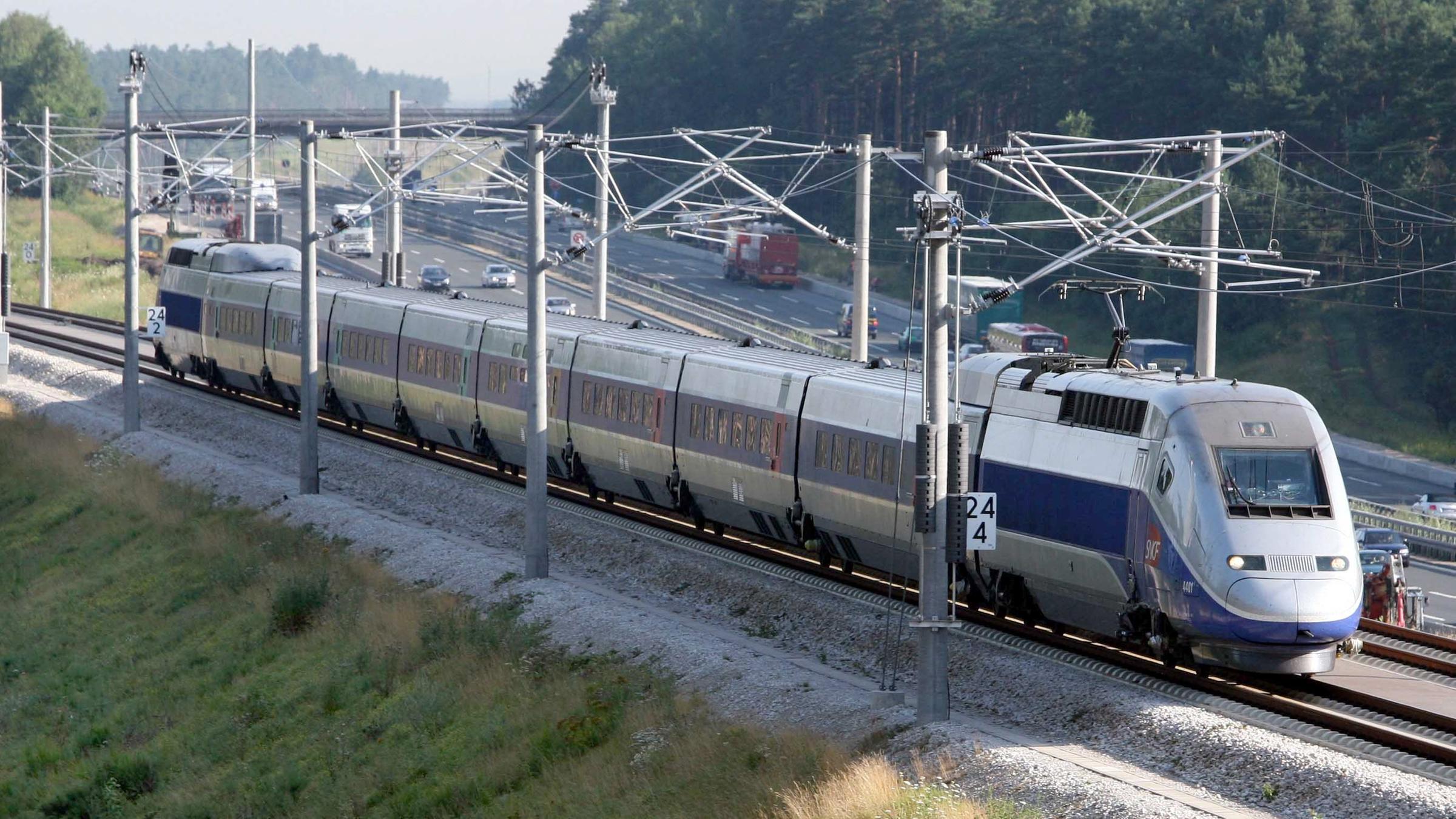 Ein TGV fährt auf einer Eisenbahnstrecke. Rechts daneben sieht man eine Autobahn.