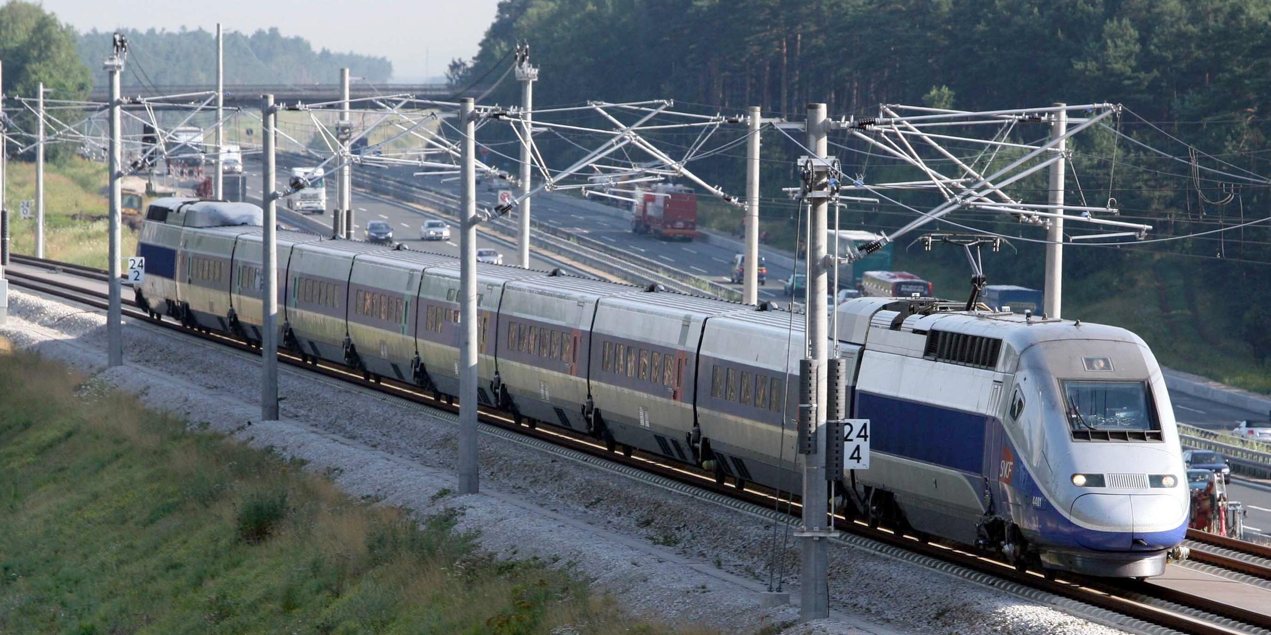Ein TGV fährt auf einer Eisenbahnstrecke. Rechts daneben sieht man eine Autobahn.