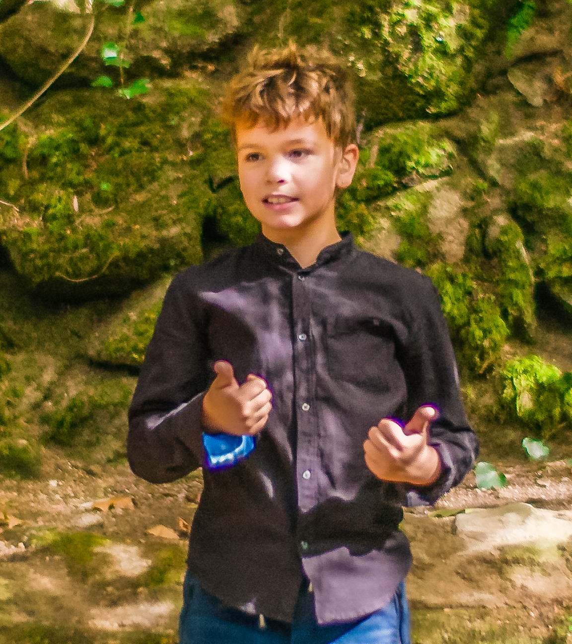 Tiago steht vor einer moosbewachsenen Felswand im Wald. Hier will er seine erste Unterrichtsstunde halten. Er trägt eine Jeans und ein dunkles Hemd und wirkt konzentriert und zuversichtlich.