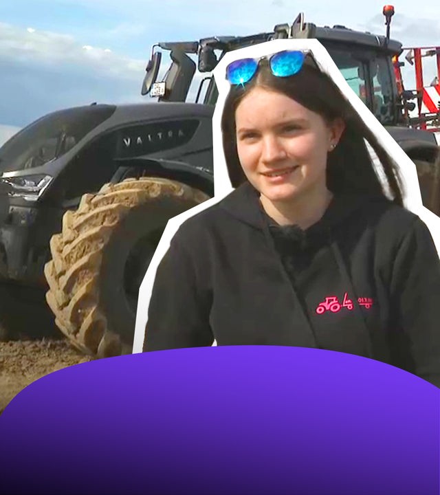 Eine junge Frau im schwarzen Pulli, im Hintergrund ein riesiger Traktor. 