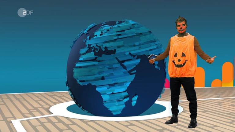 Tim Schreder ist als Kürbis verkleidet: mit Kürbisschminke und einem orangenen Kürbis-T-Shirt. Er steht im logo!-Studio