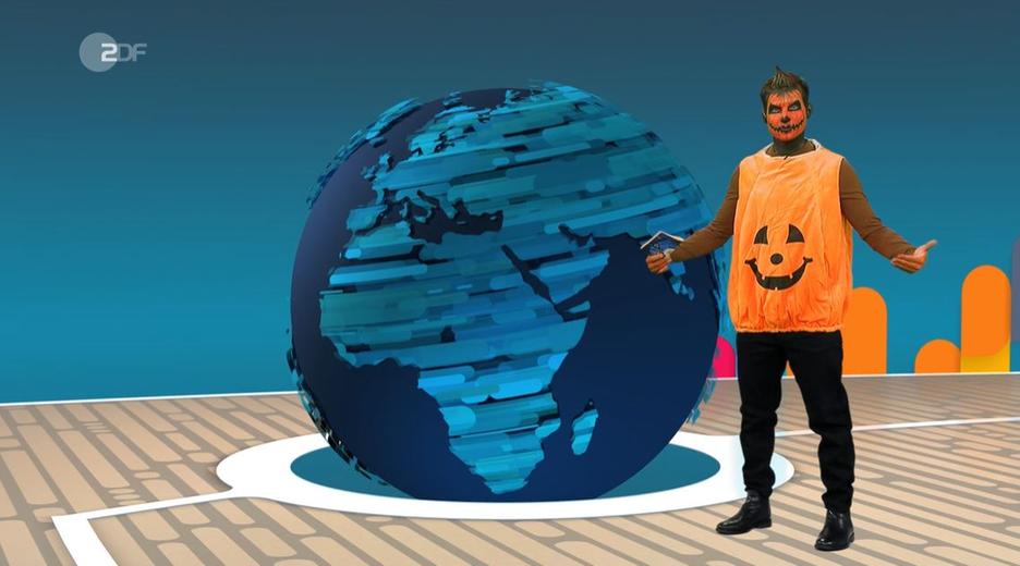 Tim Schreder ist als Kürbis verkleidet: mit Kürbisschminke und einem orangenen Kürbis-T-Shirt. Er steht im logo!-Studio