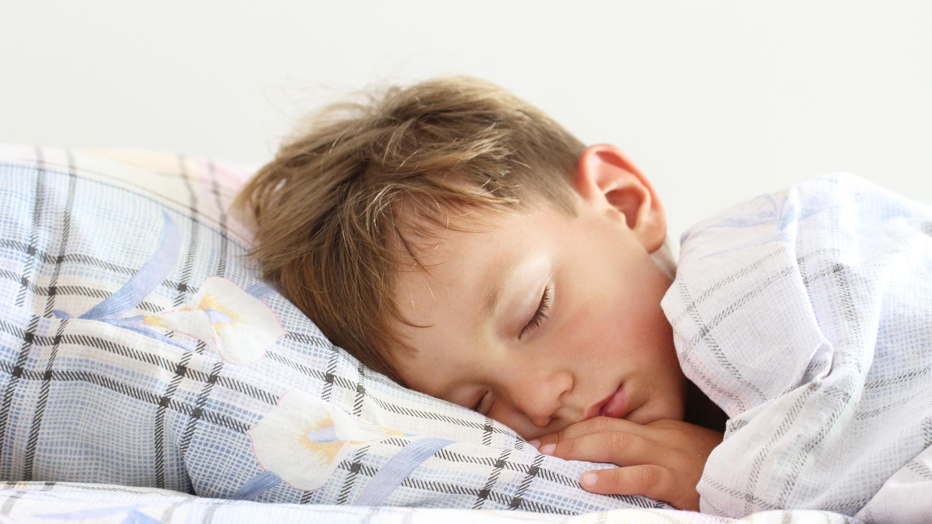 Auf dem Foto sieht man einen Jungen, der im Bett liegt und schläft. Seine Bettwäsche ist hellblau kariert, mit weißem Blumenmuster drauf.